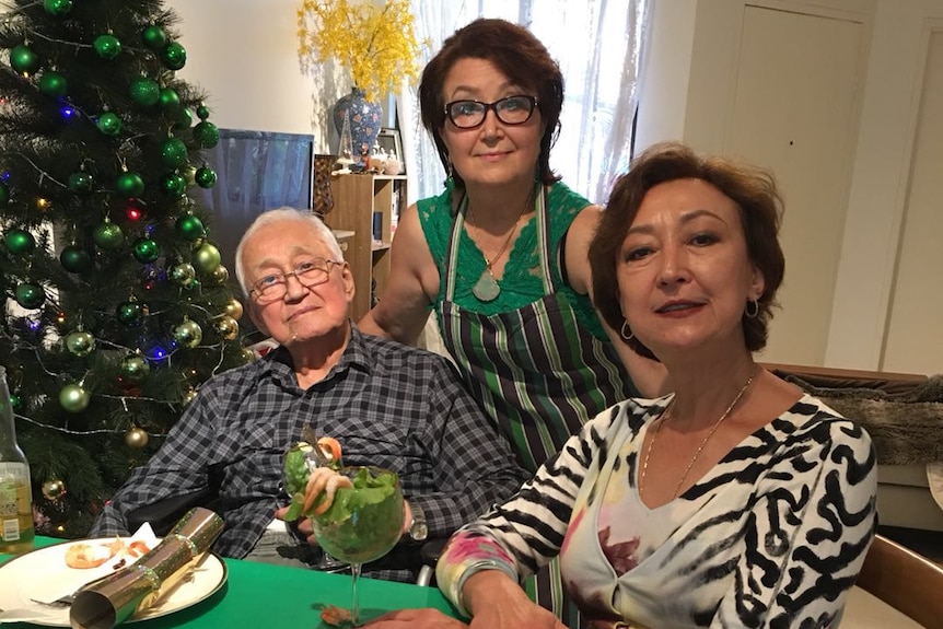 An older man sits at a Christmas lunch table with two women and a decorated tree behind them