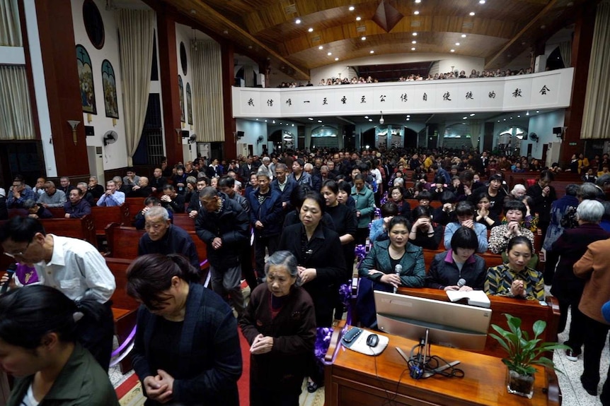 A crowd is made from people dressed in semi-formal wear bowing their heads with their hands pressed together