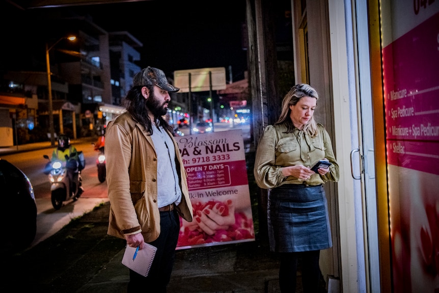 Outside a shop with pink and white branding, a woman talks to another women in a mask, as a man watches