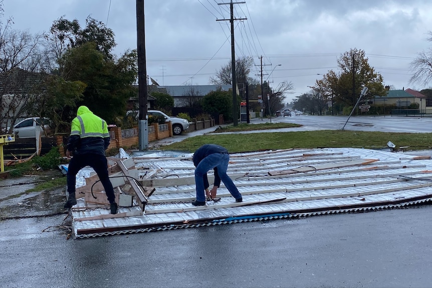 Two men work to tie chains to a metal roof laying flat on the ground.