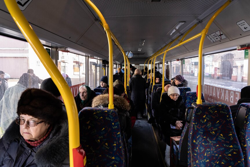 People in beanies and jackets sit on a bus with patterned seats and yellow handrails