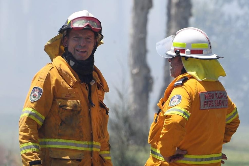 Tony Abbott, wearing protective fire gear, stands with another firefighter. There is ash all over his uniform.