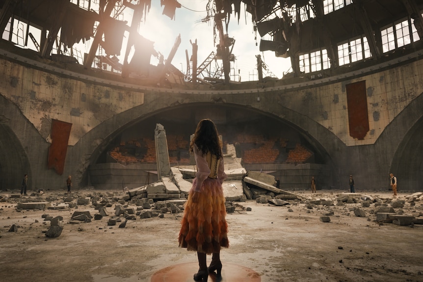 A circular arena with a giant hole in the roof, with character Lucy Gray facing the rubble.