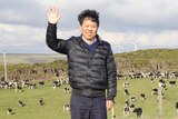 A man in a puffer jacket stands in front of the rolling hills of a dairy farm, waving.