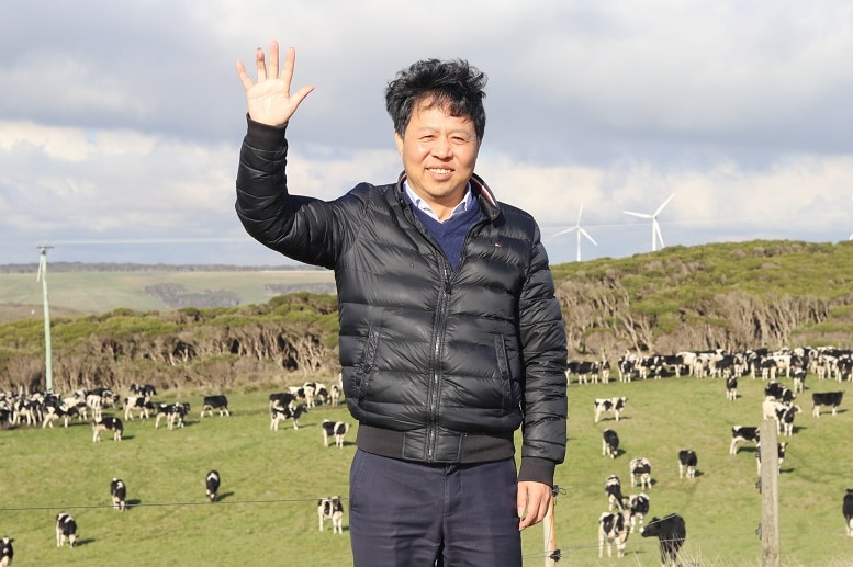 A man in a puffer jacket stands in front of the rolling hills of a dairy farm, waving.