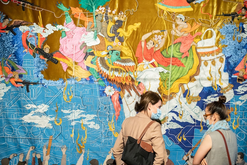 Two women in masks standing in front of a tapestry by Khadim Ali depicting mythical heroes at war