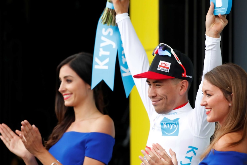 Caleb Ewan, in cycling gear and a Lotto Soudal branded cap, raises both hands on a podium next to two women.