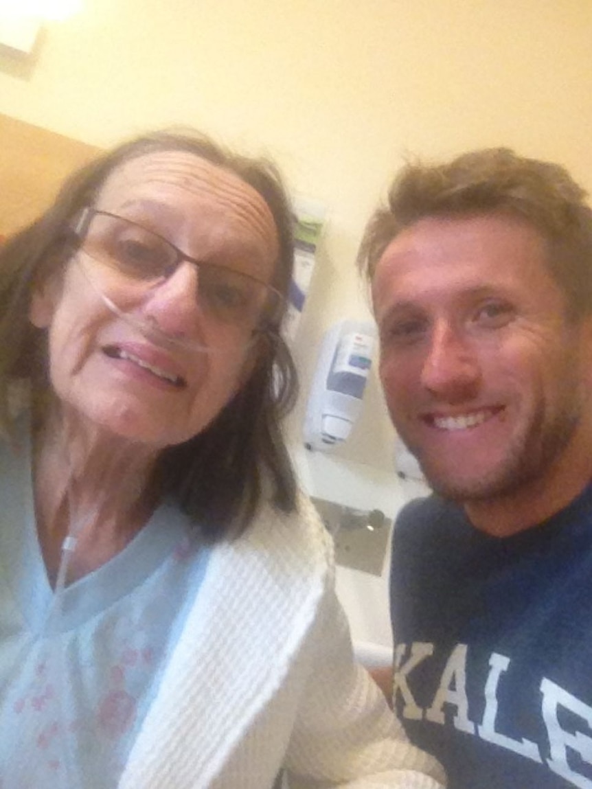 An older woman in a hospital gown, with a glasses and a tube in her nose, next to a younger man. Both smiling.