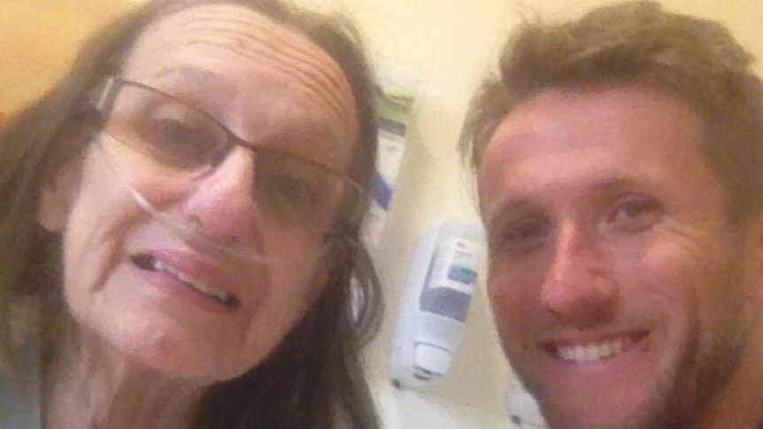 An older woman in a hospital gown, with a glasses and a tube in her nose, next to a younger man. Both smiling.