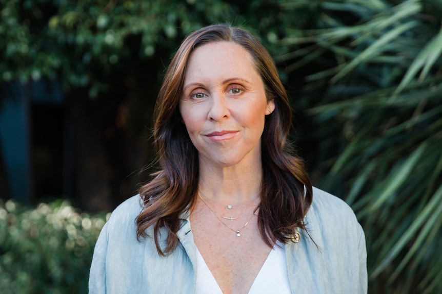 Woman has brown wavy hair, looks front on at camera with white top and grey jacket, green palms in background