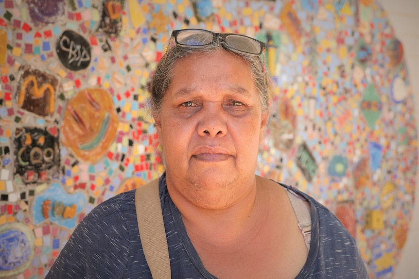 An older woman, grey hair tied, glasses on top, in front of a colourful wall with Indigenous art.