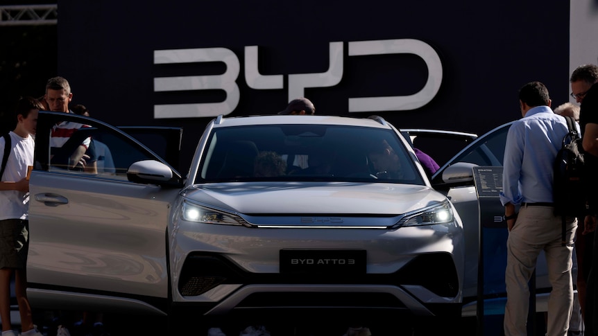 几个人围在一辆白色汽车旁观看，汽车后面的墙上有BYD三个字母。