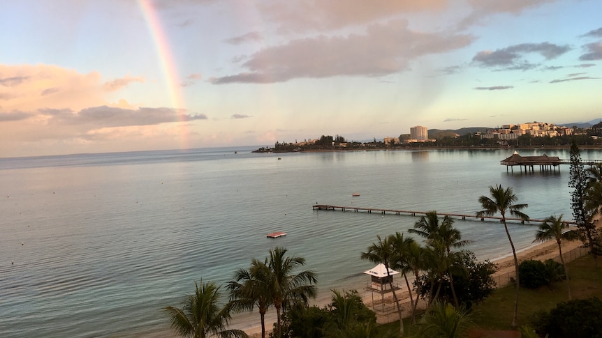 A pontoon juts into a calm bay over which a rainbow arcs, with a few tall buildings in the background.