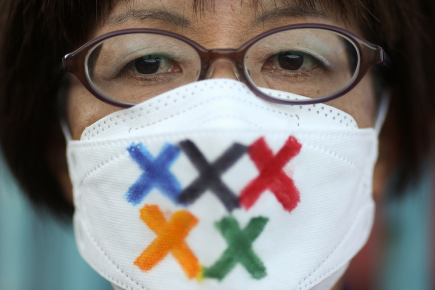 A close-up of a Japanese woman wearing a mask in protest with the Olympic rings painted on as crosses. 