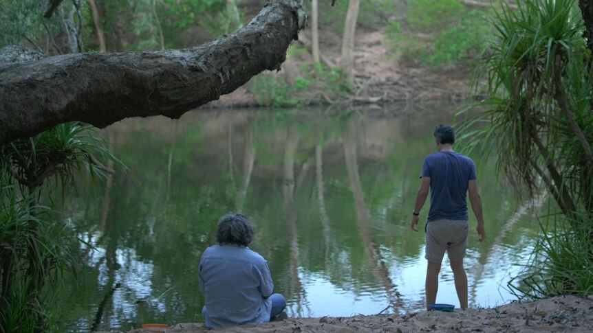 One man stands and another kneels with their backs turned while looking over a creek