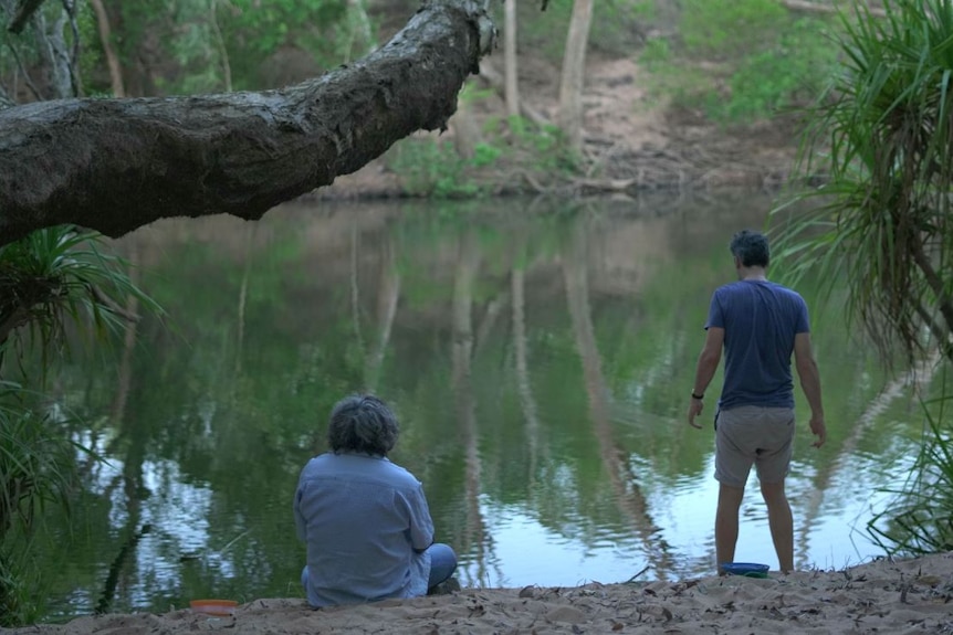 One man stands and another kneels with their backs turned while looking over a creek