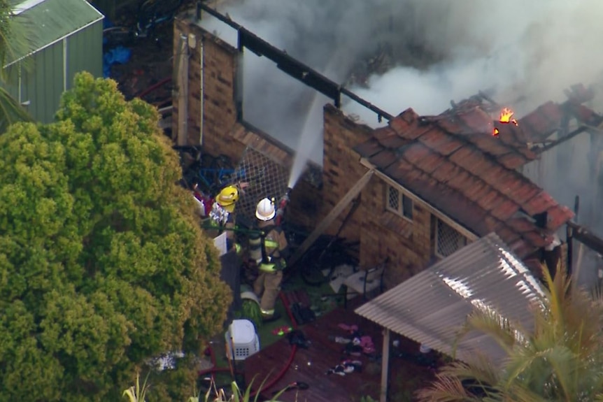 Two firefighters outside a burning house, one holding a hose. The roof has collapsed.