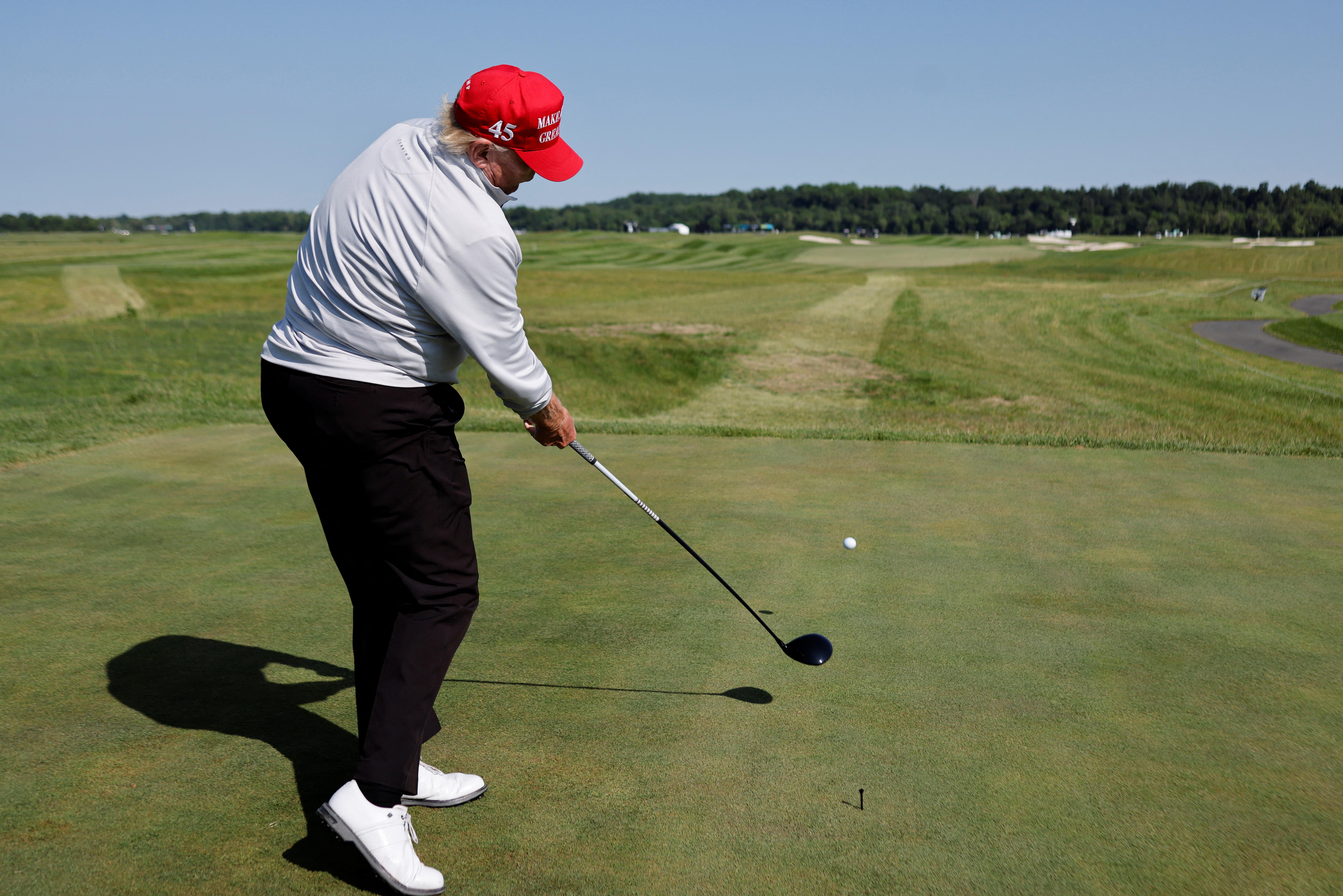  Donald Trump wears a red MAGA cap and white shirt as he hits a tee shot in an open golf course.