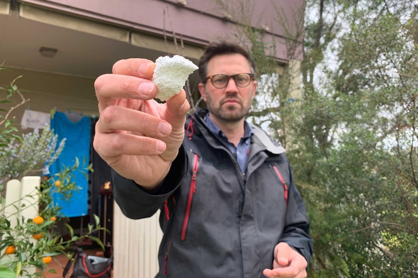 A man standing in front of an apartment holding a piece of polystyrene foam towards the camera.