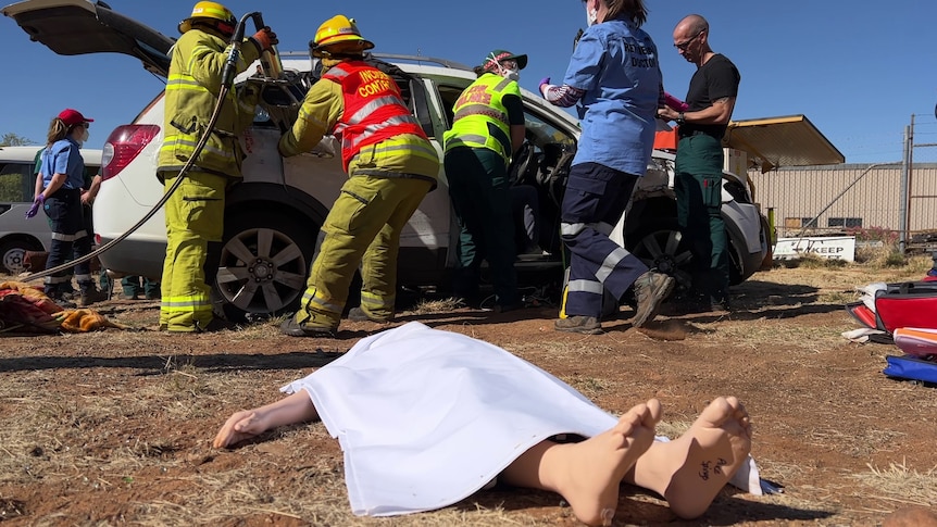 Dummy under a white sheet in the foreground, firefighters and medical staff work around a white car in the background.