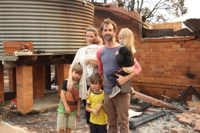 A family stand in front of a destroyed house.