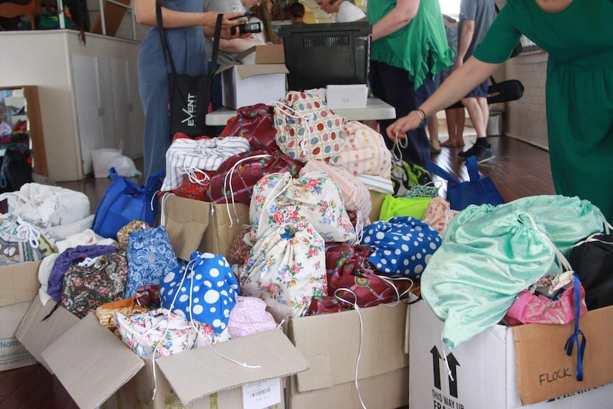 Bags are seen sitting in boxes at a CWA hall. People are seen behind the boxes packing more bags.