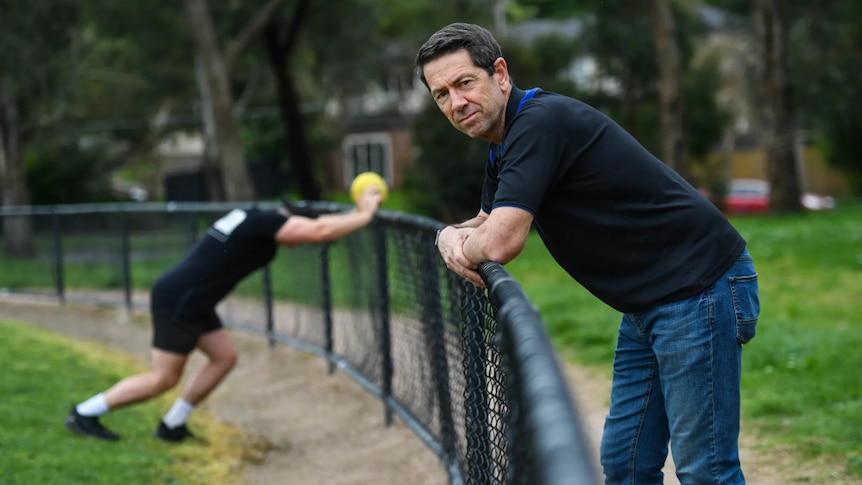 man wearing jeans and dark top leans on football oval fence. person stretching in background