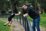 man wearing jeans and dark top leans on football oval fence. person stretching in background