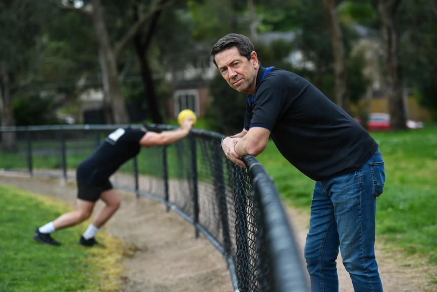 man wearing jeans and dark top leans on football oval fence. person stretching in background