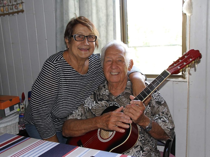 A man sits, holding a guitar, while his wife leans on his shoulder.
