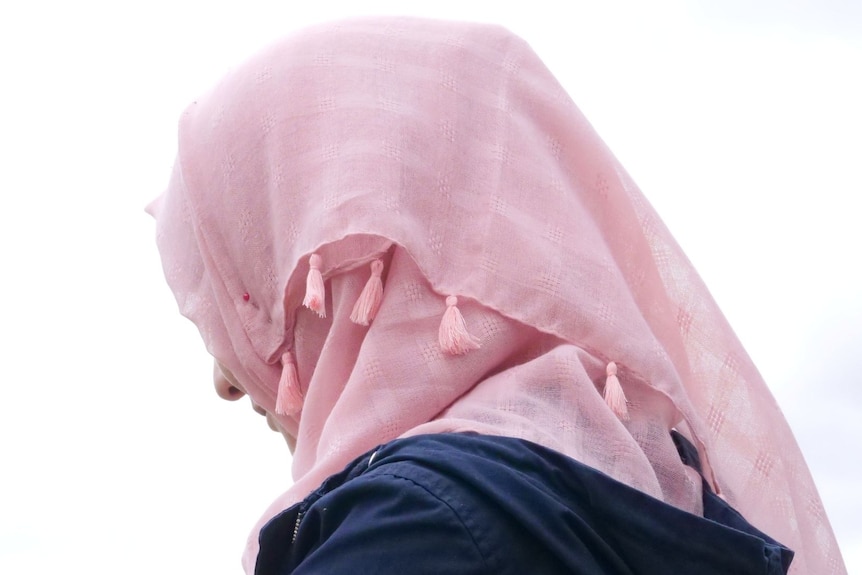 A woman wearing a pink hijab looks away from the camera outdoors. 