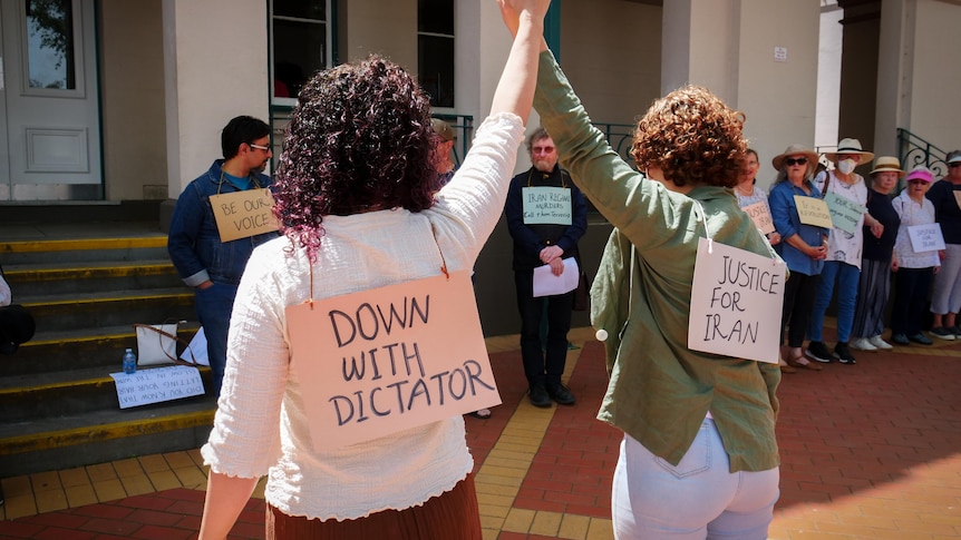 Two women stand facing a group of protesters wearing signs seeking justice for Iran 