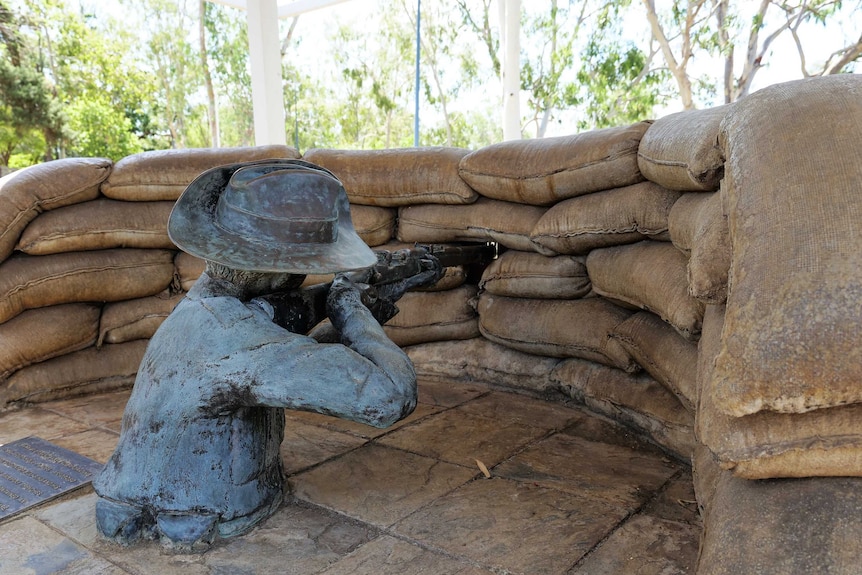 a statue of a man holding a rifle and aiming it through a whole in a barrier created by sandbags