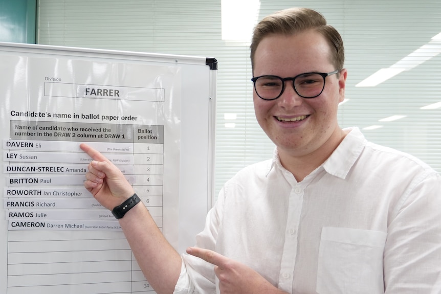 A young man in a white shirt and glasses points to his name on a voting ballot that says Farrer