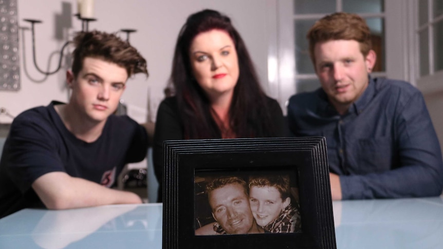 Three family members sit behind photo of the late father.