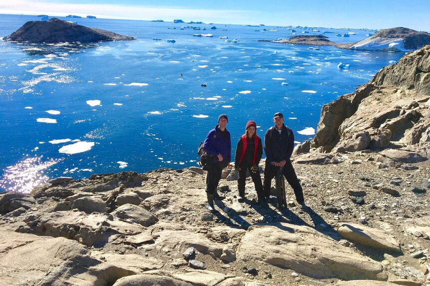 Three people stand on an island, with an ocean and icebergs in the background.