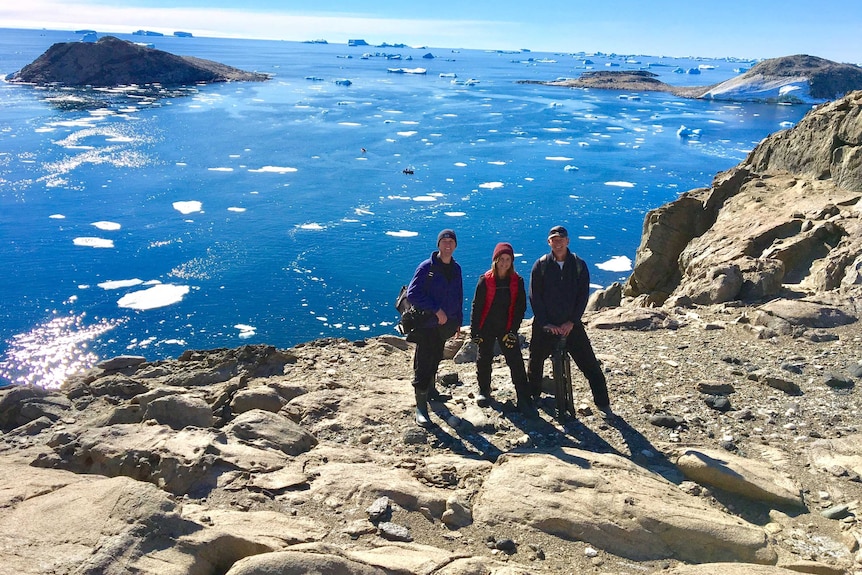 Three people stand on an island, with an ocean and icebergs in the background.