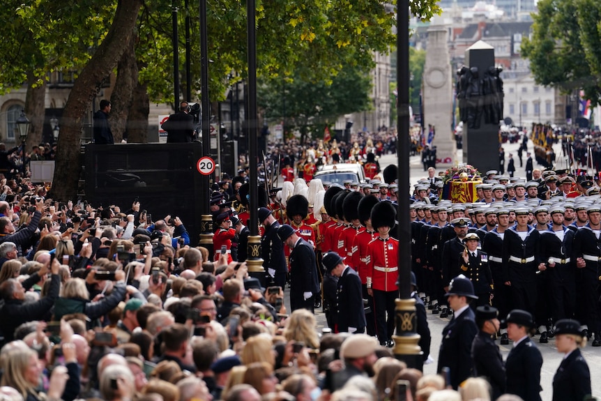 On the left a large crowd of people is watching the Queen's procession on the right pass by.