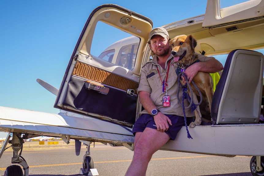 A man's hand is around the dog.  They are both standing in front of a small plane. 