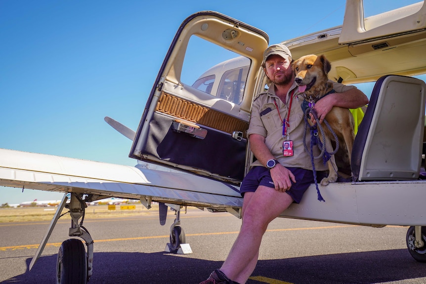 A man has his arm around a dog. They both stand in front of a small plane. 