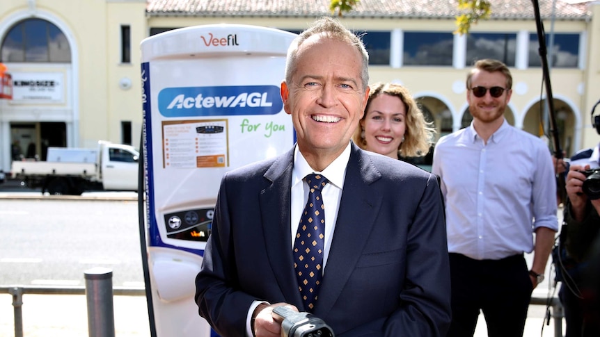 Bill Shorten smiles as he looks at the camera holding an electric car charger