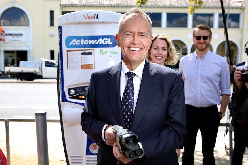 Bill Shorten smiles as he looks at the camera holding an electric car charger