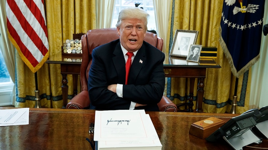 Donald Trump sits at his desk in the Oval Office with his arms crossed.