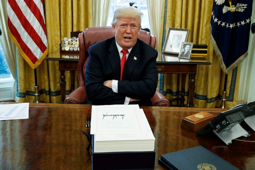 Donald Trump sits at his desk in the Oval Office with his arms crossed.