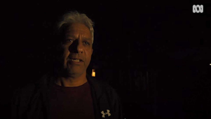 An Indigenous man against a black background at night talks by the flickering light of camp fire.