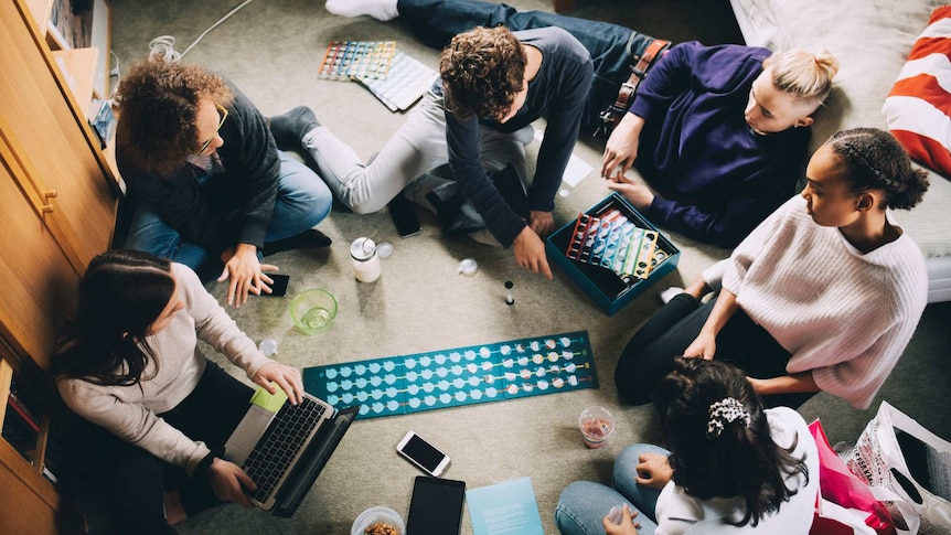 top view of a group of young adults sitting in a bedroom, looking intently at a boardgame in the centre of the room
