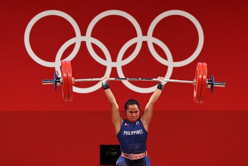 A female weightlifter from the Philippines lifts weights above her head, the Olympic rings are in the background.