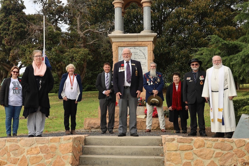 A group of people, some wearing medals and poppies, stand in front of a memorial.