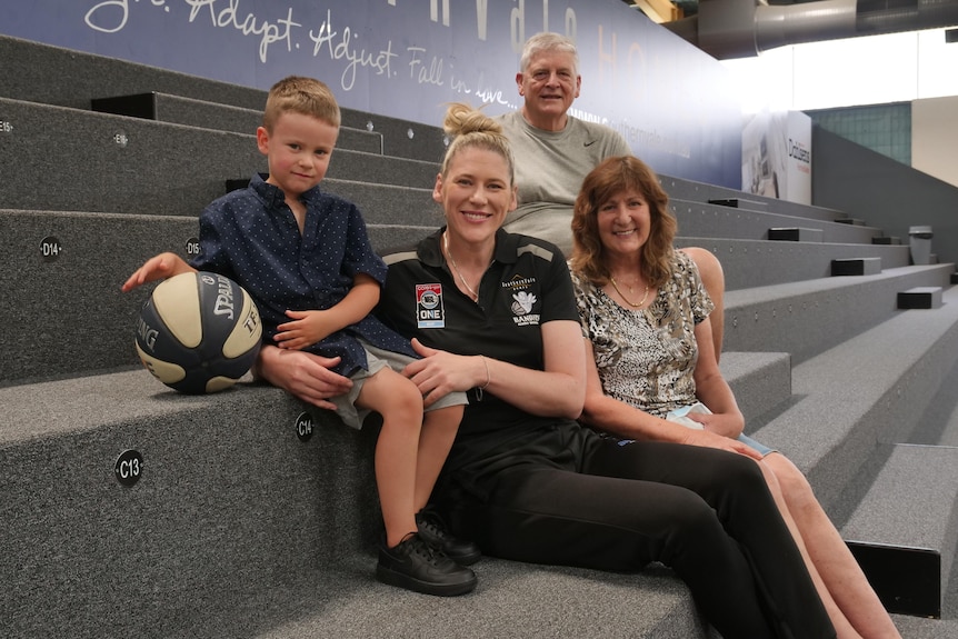 young boy sits on stadium grandstand holding basketball next to woman and older couple