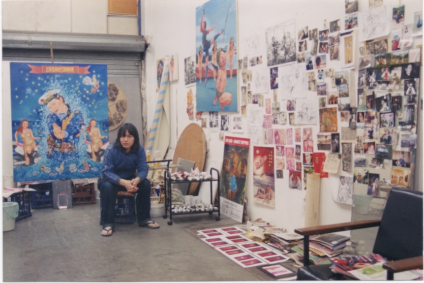 A young Chinese man sitting in a studio with paintings and artworks.